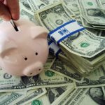 Stock art of a bunch of money with a piggy bank in front and a hand putting a coin in