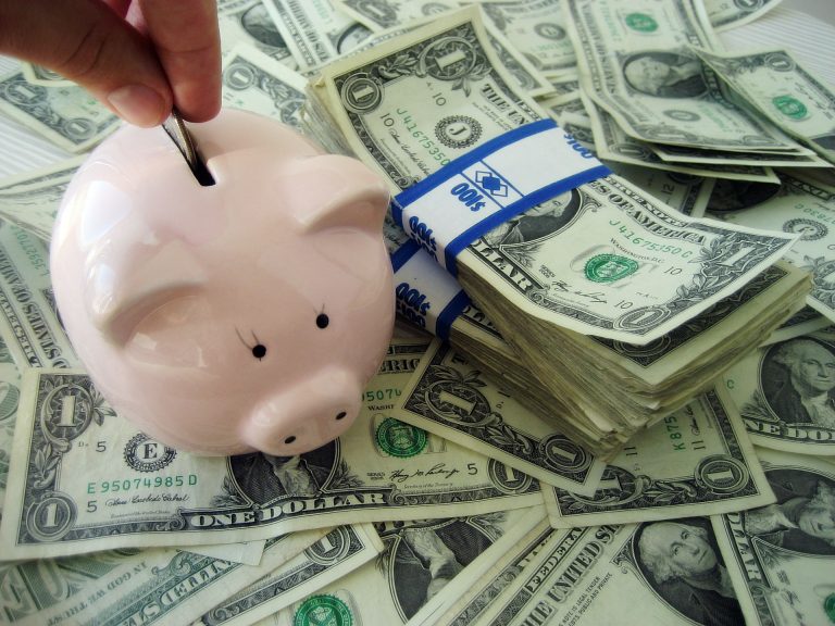 Stock art of a bunch of money with a piggy bank in front and a hand putting a coin in