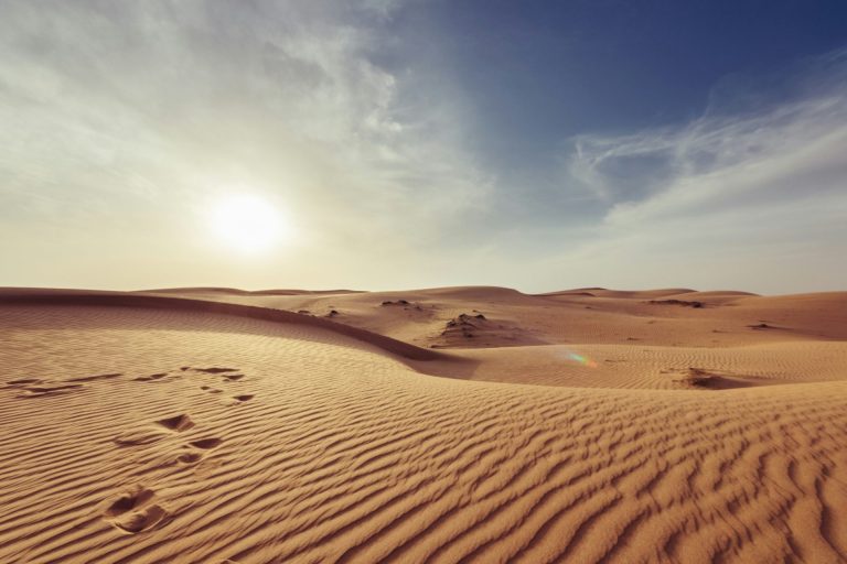Footprints disappearing in a sparse, hard desert landscape with the sun blazing in the afternoon sky.