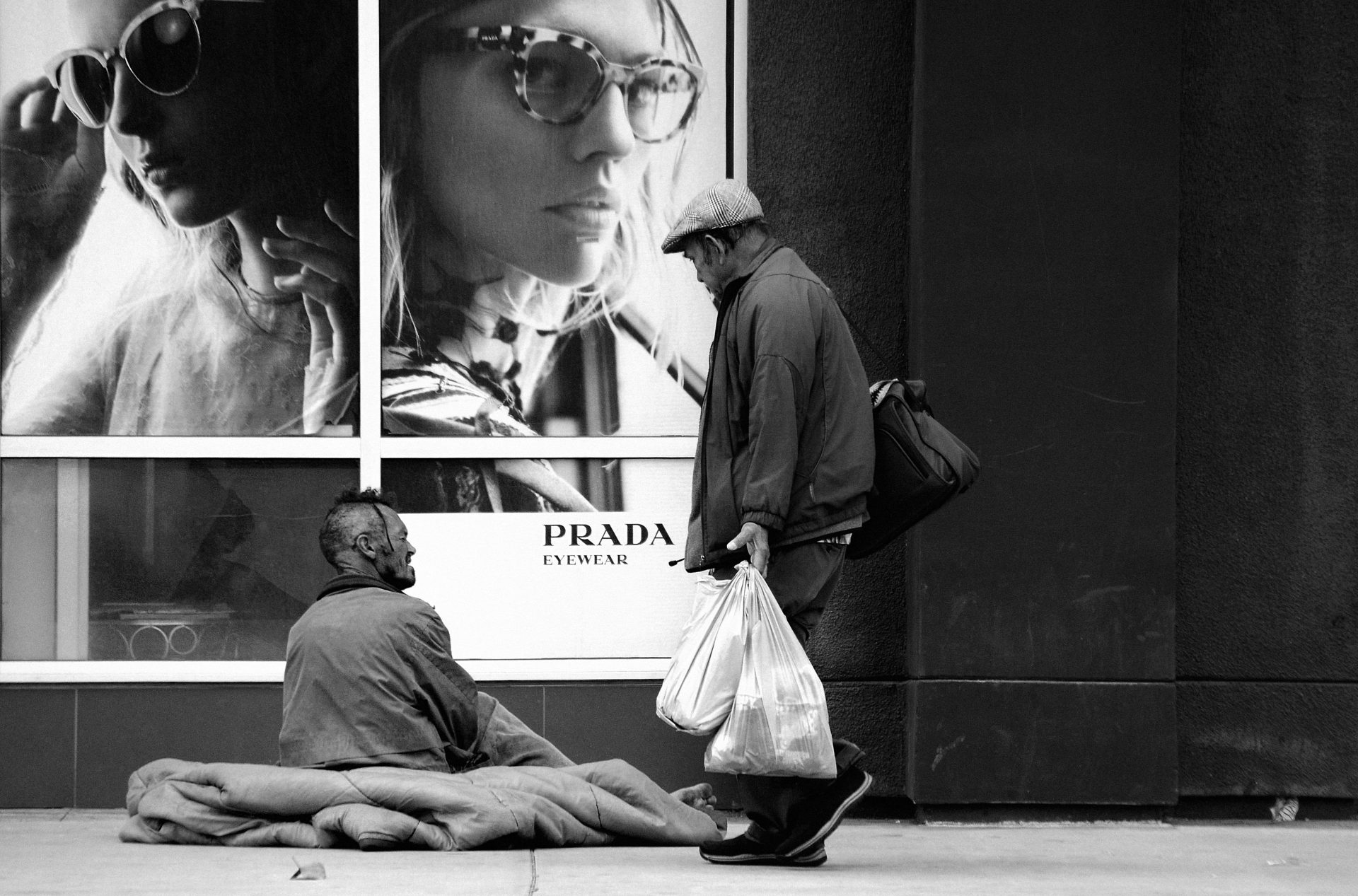 Homeless person sitting on side of building with his sleeping bag in front of a PRADA fashion sign