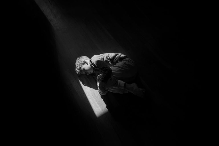 Noir highly styled photograph of a boy lying on a wooden floor the camera looking down on him from above with light coming from a small skylight window above
