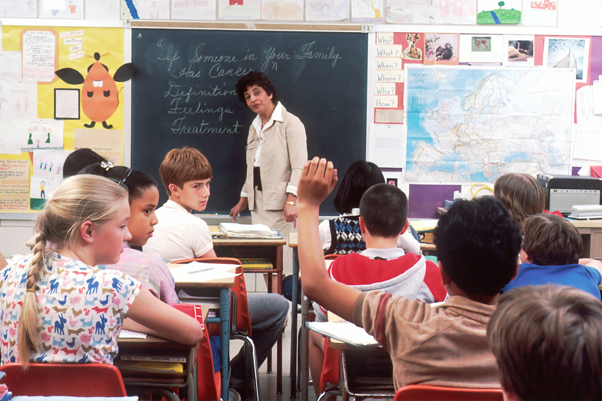 A group of middle school kids and teacher looking at a student who has their hand raised to ask a question
