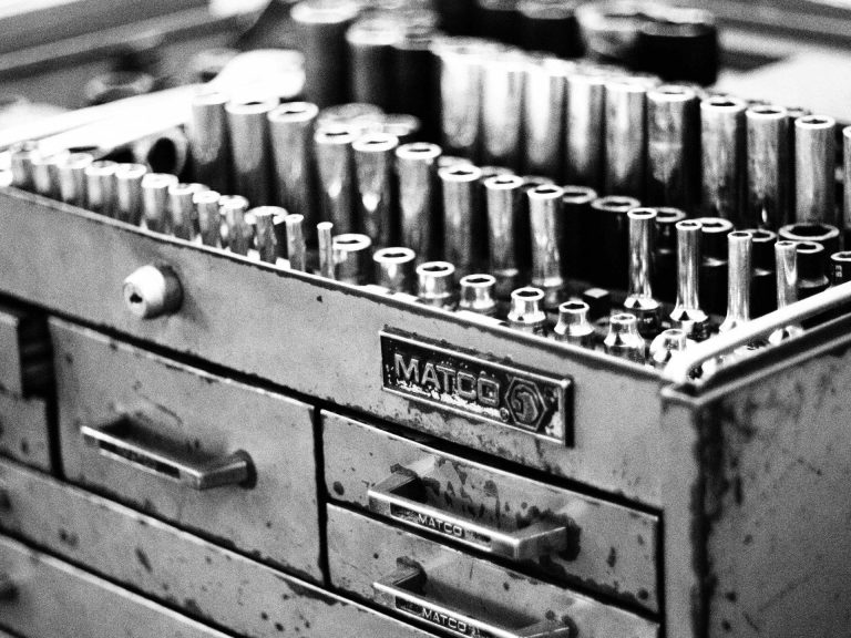 CLoseup black and white photo of a roughly used toolbox featuring a sharp-looking socket set in the open top shelf.