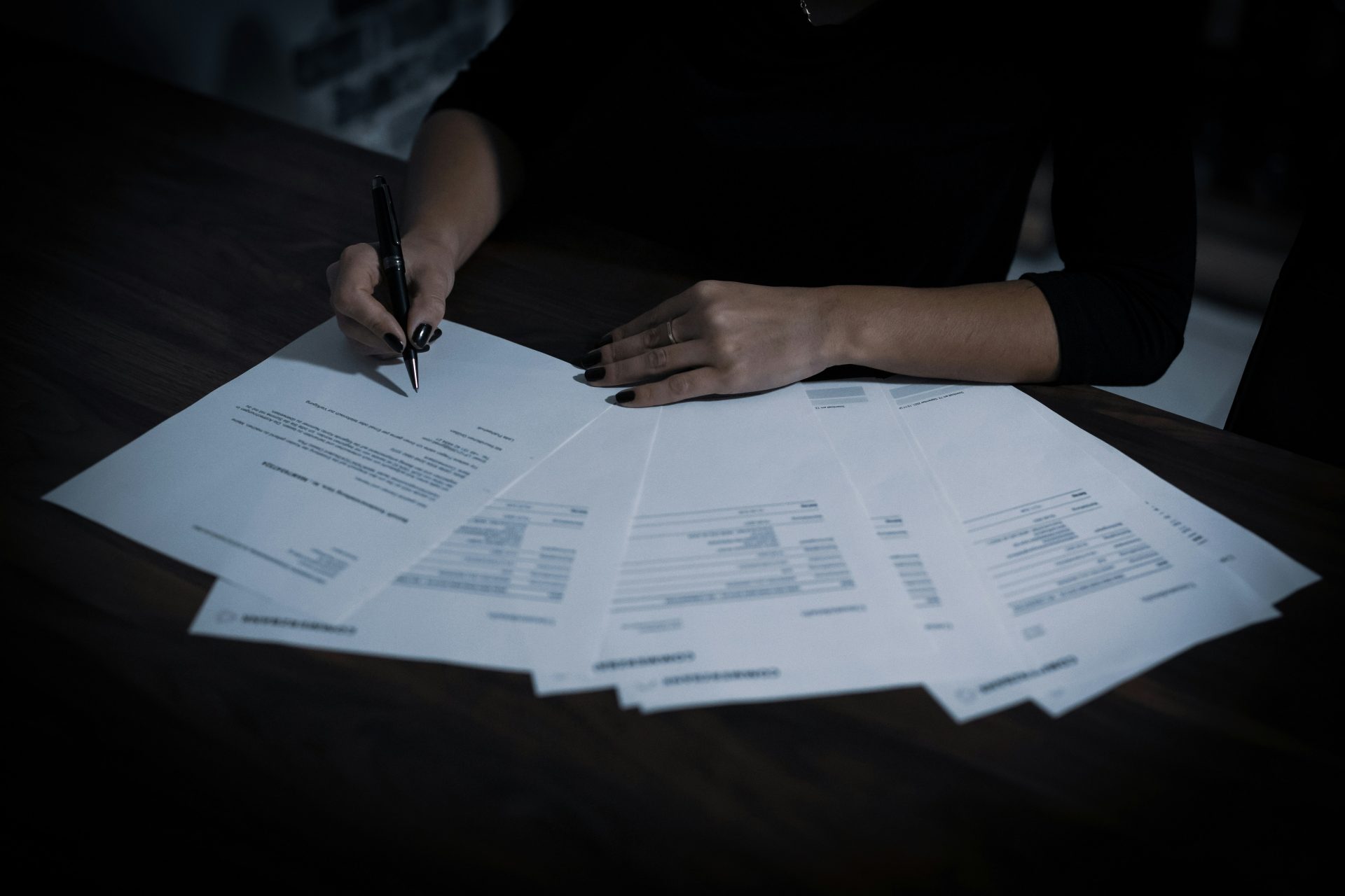 A spread of contract paperwork and a pair arms and hands reviewing and signing them... everything else in darkness.