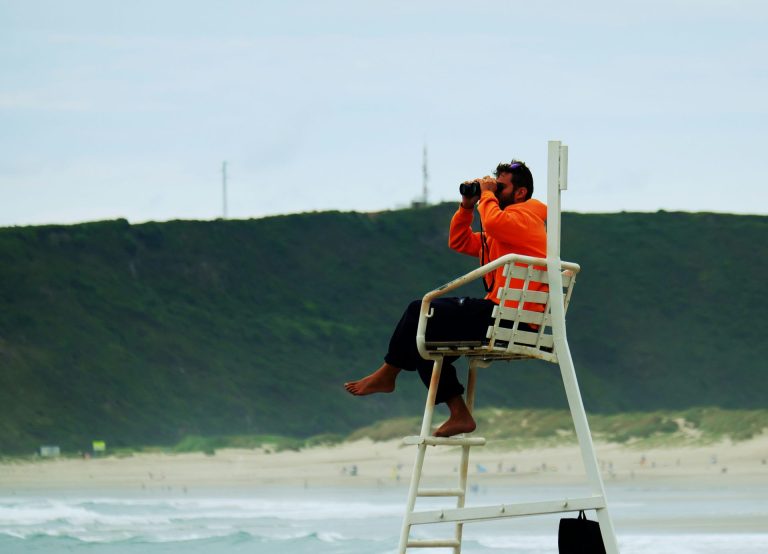 Lifeguard sitting in his high chair looking out into the surf with binoculars.