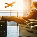 Young guy sitting in an airport departure area with his feet up on his carryon looking out at a plane taking off