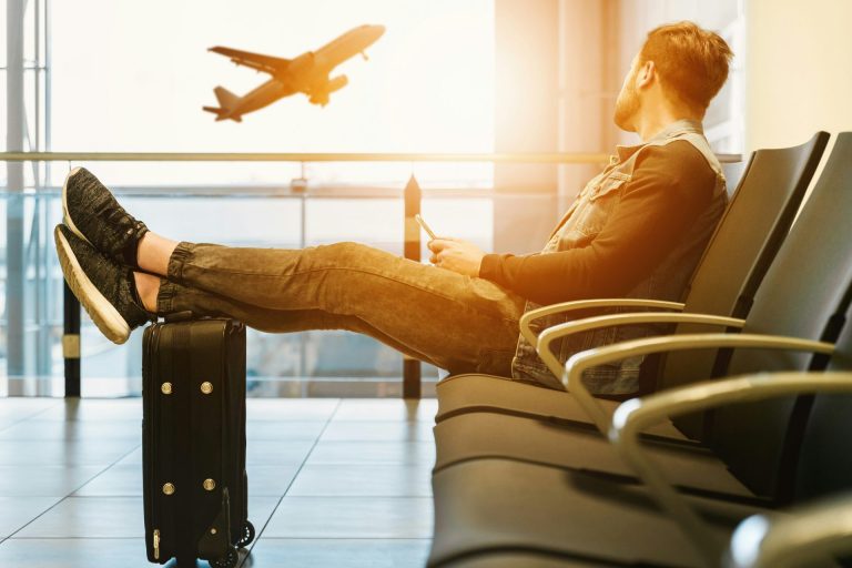 Young guy sitting in an airport departure area with his feet up on his carryon looking out at a plane taking off