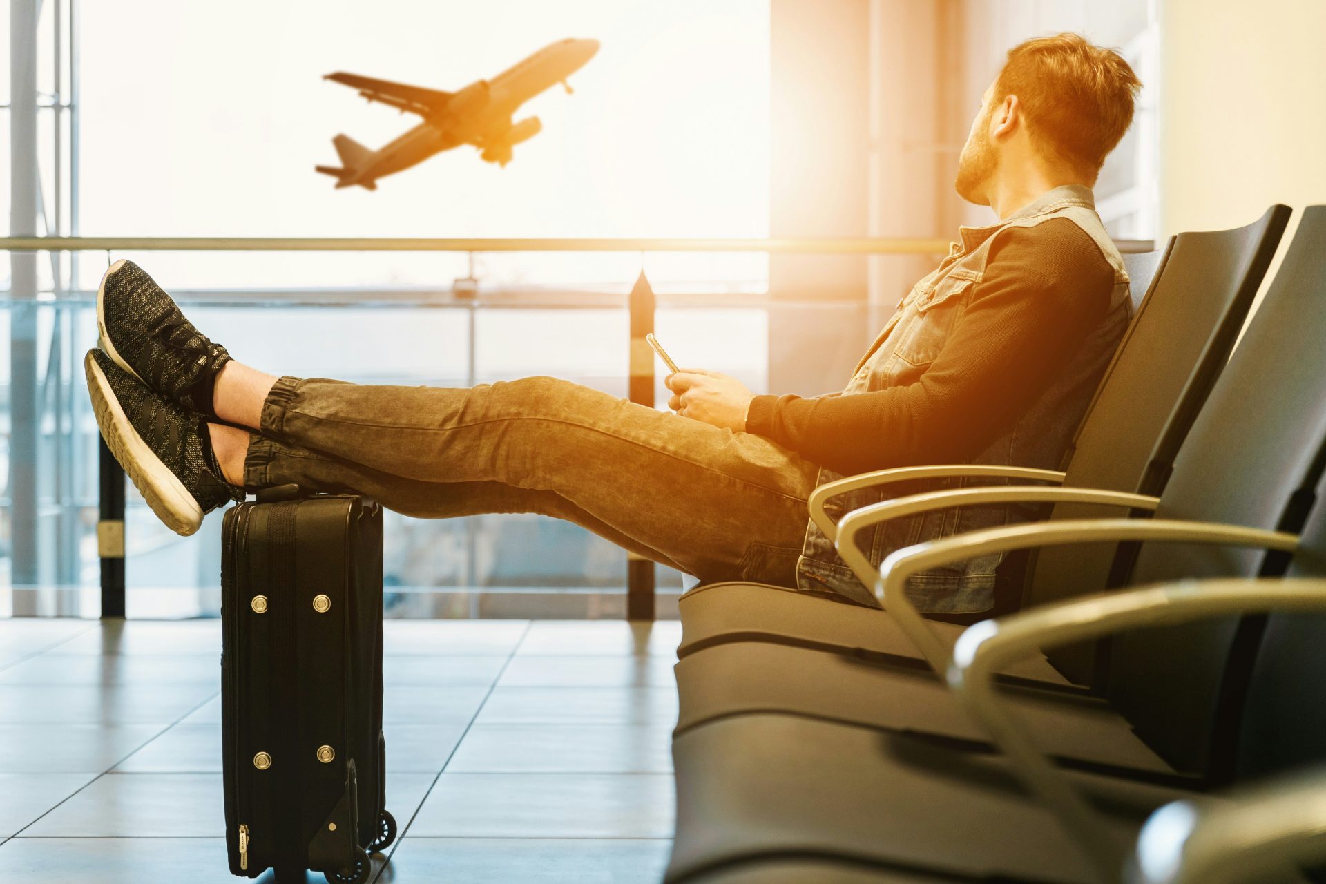 Young guy sitting in an airport departure area with his feet up on his carryon looking out at a plane taking off