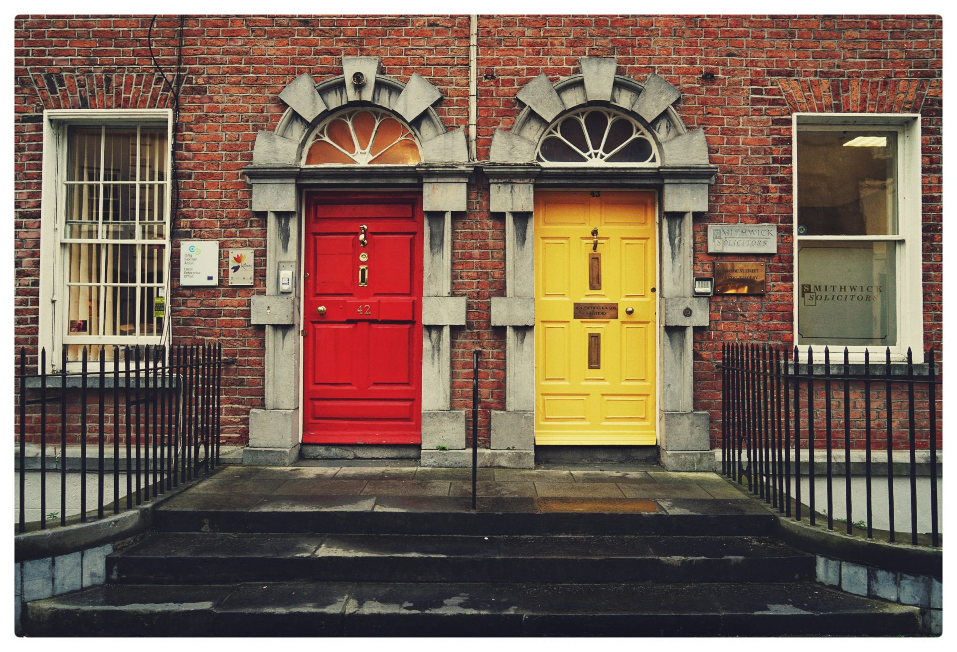 A 19th century brick duplex building with two brightly colored doors - one red and one yellw.