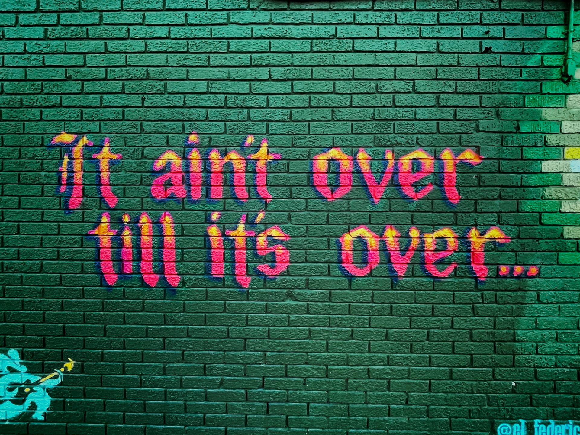 Green brick wall with Orange and Red Calligraphic Graffiti saying "It ain't over till it's over..."