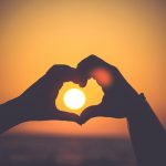 Silhouetted hands making a heart gesture with the sun at sunset behind the heart.