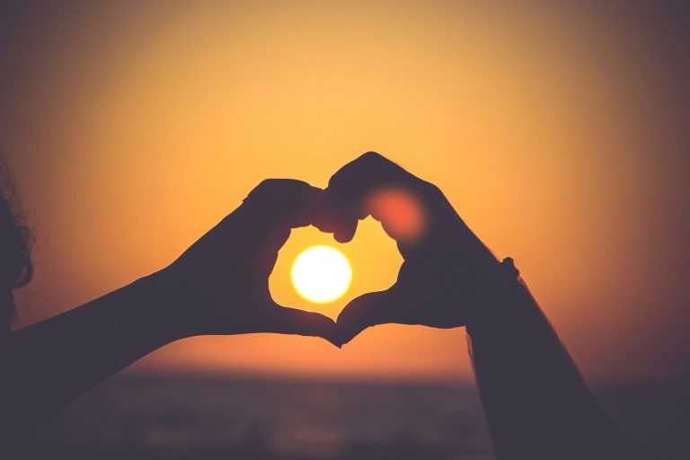 Silhouetted hands making a heart gesture with the sun at sunset behind the heart.